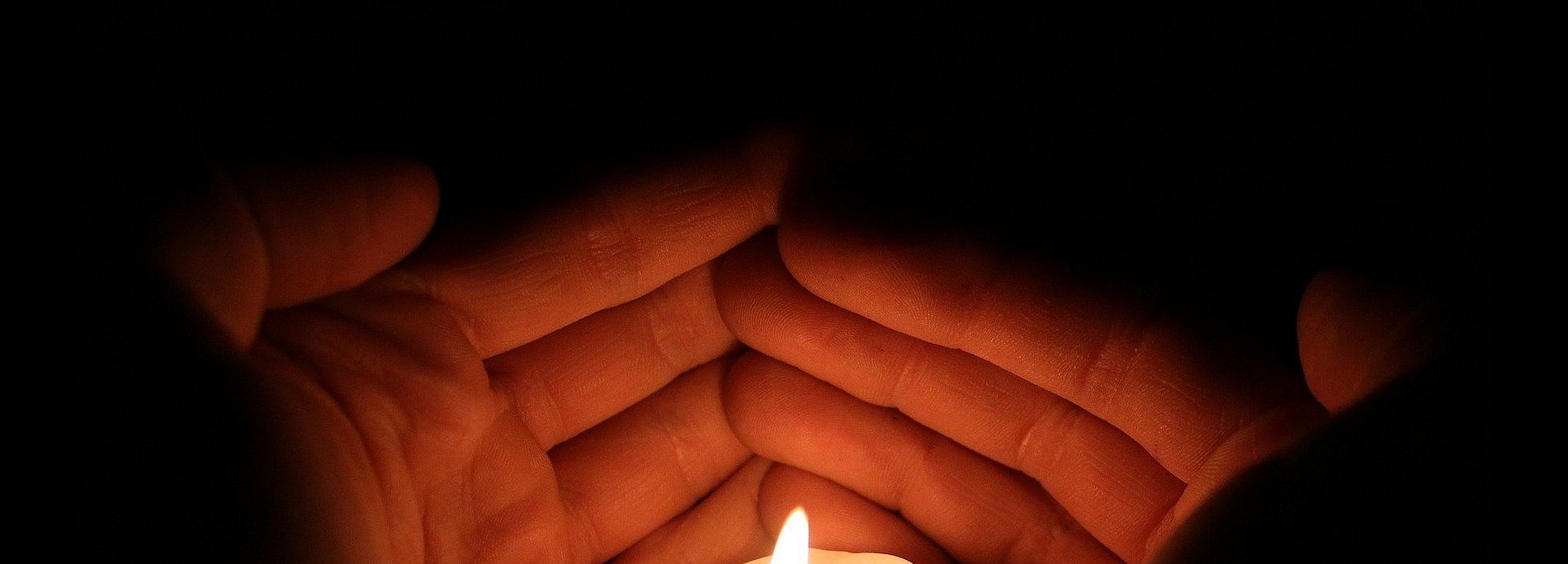 Zwei Hände, die um eine brennende, weiße Stumpenkerze gelegt sind, wie um die Flamme zu schützen. Schwarzer Hintergrund.